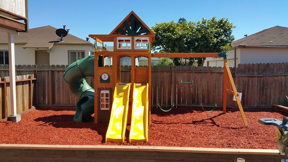 Backyard play structure with slides and swings installed inside a fenced yard.